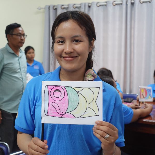 A teacher smiles and holds up a coloured fish-shaped craft at a workshop