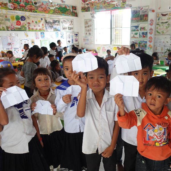 Children hold up their folded paper creations in a crowded classroom