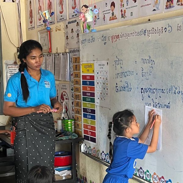 A TukTuk staff member watches a girl write on the classroom whiteboard