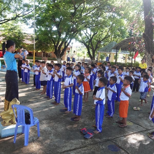 Preschool children in blue and white uniforms stand in rows outdoors while a teacher leads