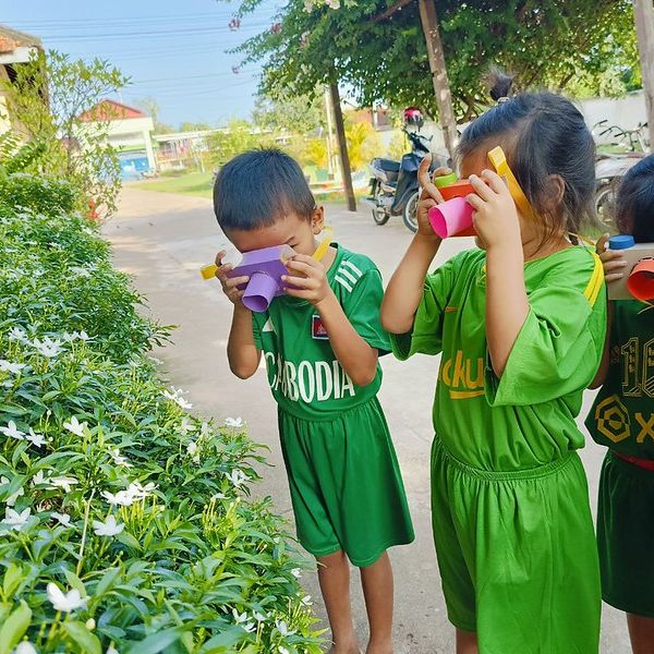 Two children in green shirts look through paper tube viewers beside a flowering hedge