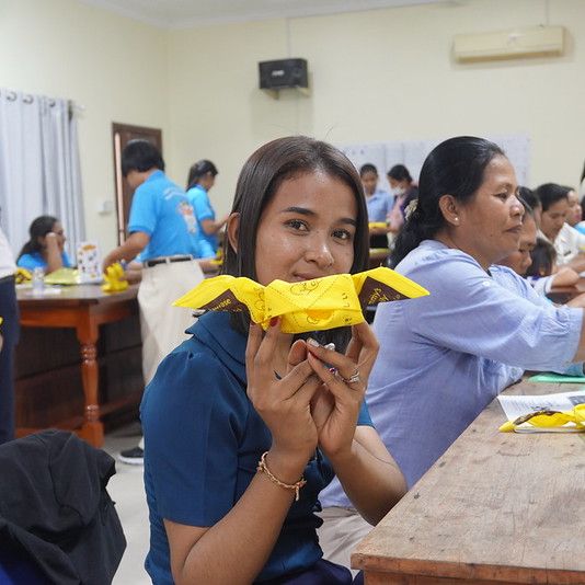 A teacher holds up a yellow folded paper craft at a workshop table