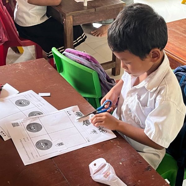 A boy cuts out a worksheet with scissors at a classroom desk