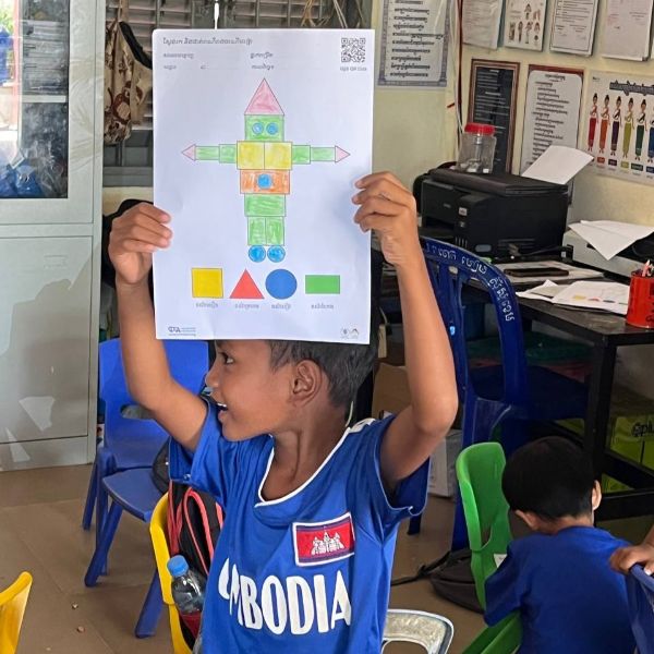 A boy holds up a coloured shape-person worksheet and smiles in class