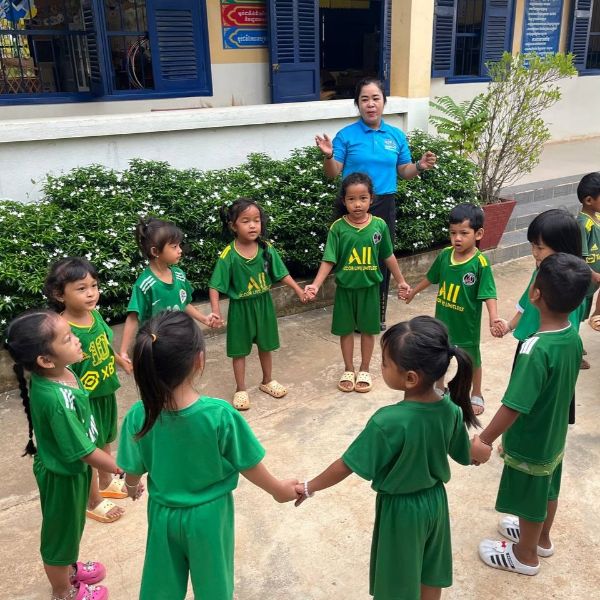 Preschool children in green hold hands in a circle outdoors while a TukTuk staff member leads