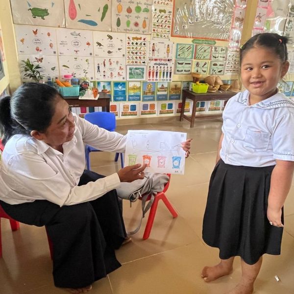 A teacher shows a girl's coloured worksheet while the girl stands smiling beside her