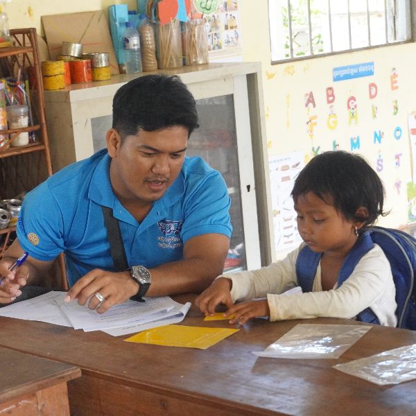 A TukTuk staff member helps a child with a craft worksheet at a school desk