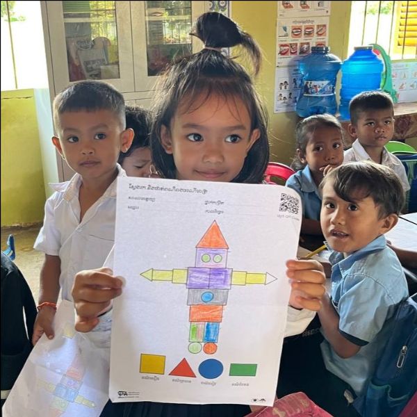 A girl holds up her coloured shape-person worksheet while classmates look on