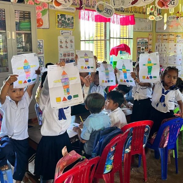 Children seated on red chairs hold up coloured shape worksheets showing robot figures in a classroom
