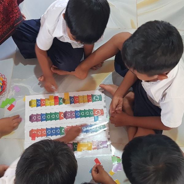 Children kneel around a floor mat sticking coloured paper pieces onto a recycling bin sorting activity sheet