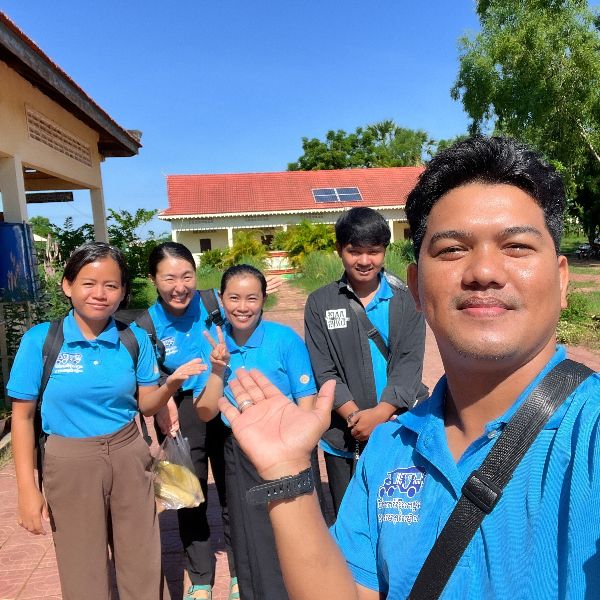 TukTuk staff in blue polo shirts smile and wave in a group selfie outside a school building