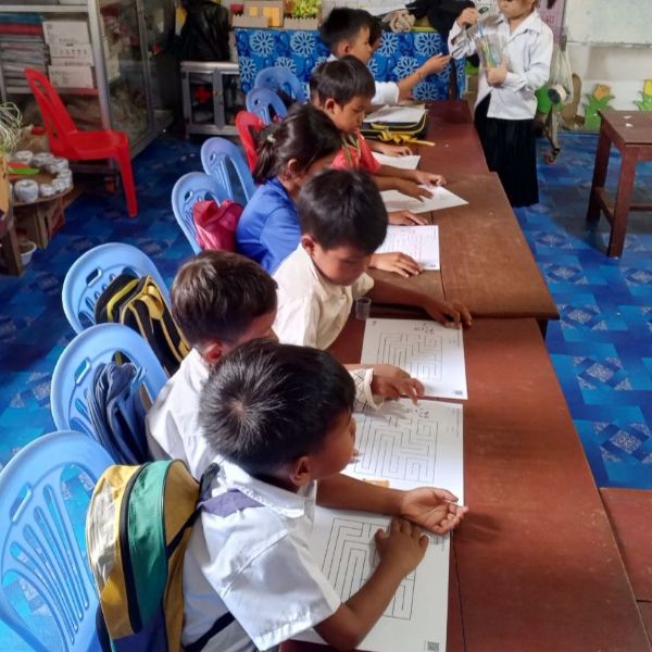 Children in school uniform sit along a long table tracing maze worksheets in a classroom