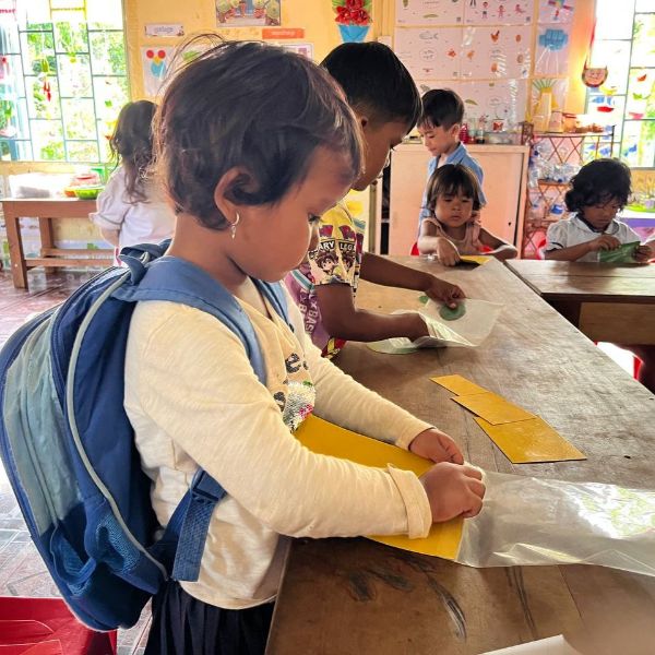A young girl with a backpack folds yellow paper at a classroom table while other children work nearby