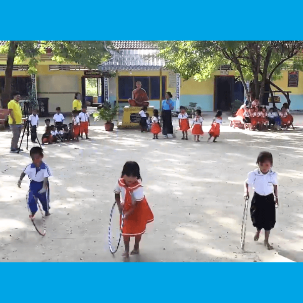 Children play with hoops in a sunny school yard while staff film and other children sit under a shelter