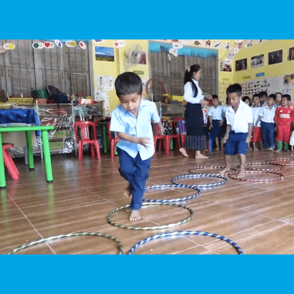 A boy hops between hula hoops laid on a classroom floor while classmates and a teacher watch