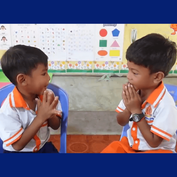 Two boys in school uniforms sit facing each other on blue chairs with their hands pressed together in greeting
