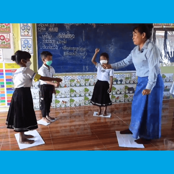 A teacher guides three masked pupils standing on paper sheets on the classroom floor in front of a blackboard