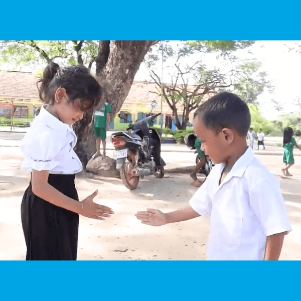 A girl and a boy in school uniforms greet each other with outstretched hands outdoors under a tree