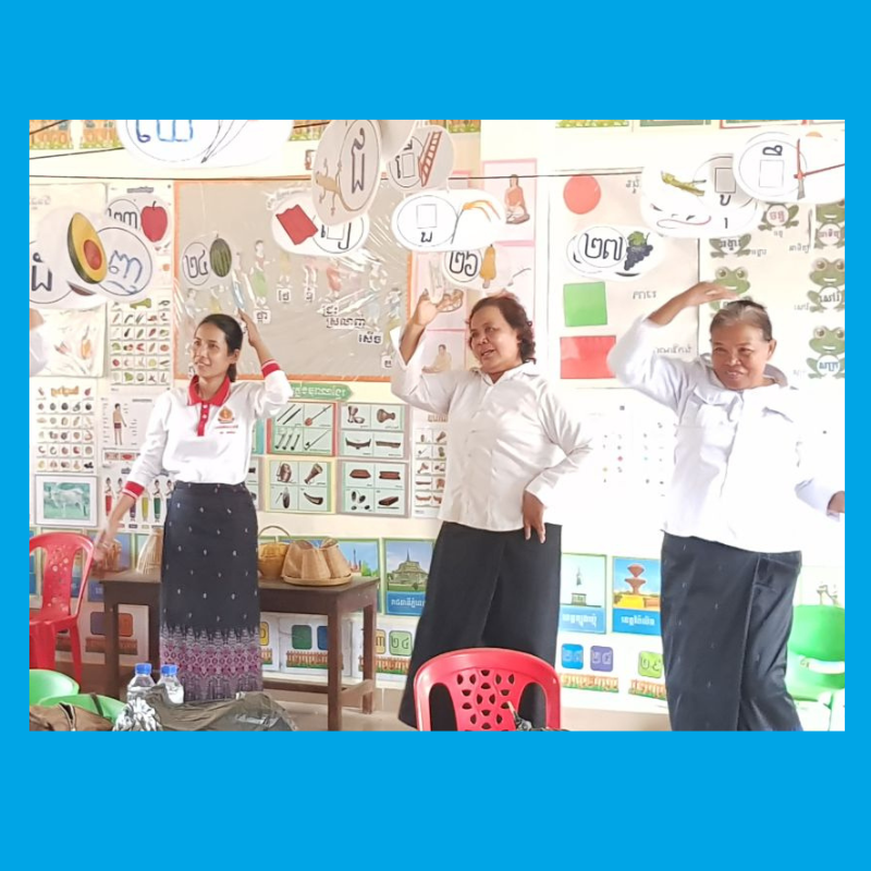 Three teachers in white tops raise their arms in a movement activity in a classroom with Khmer letter charts