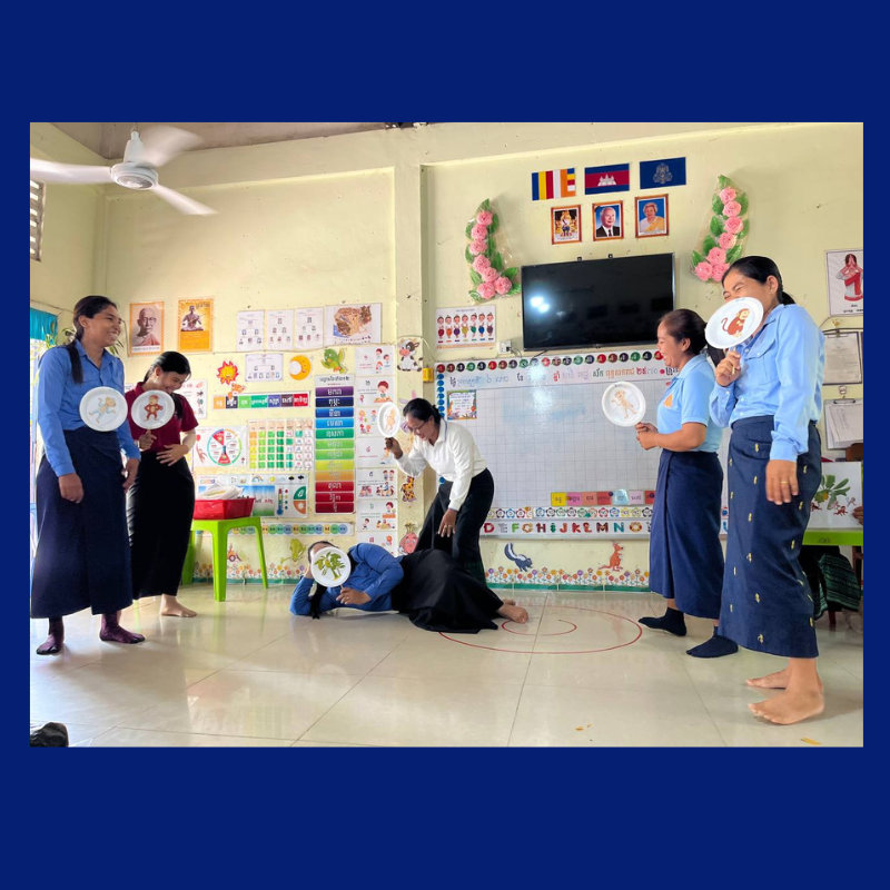 Teachers in a classroom act out a role play holding round picture cards, in front of learning charts