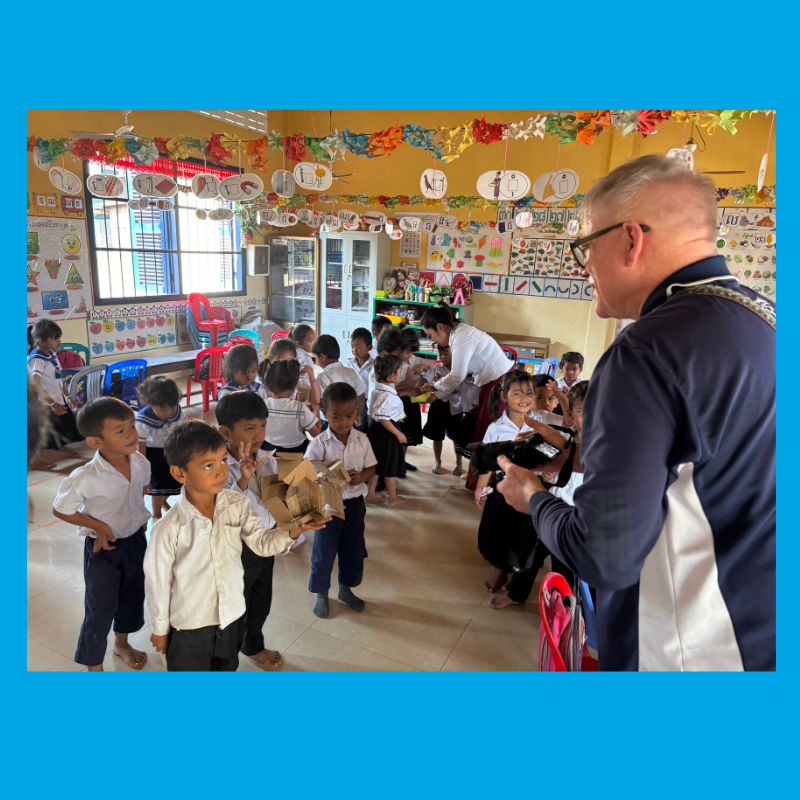 A visiting man in a navy jacket faces a crowded preschool classroom of children in white shirts