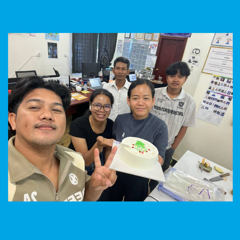 TukTuk staff gather in an office for a selfie, one holding up a decorated white cake