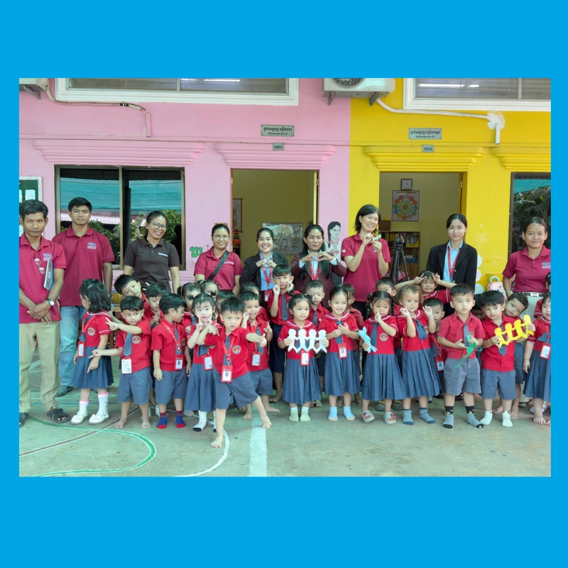Preschool children in red shirts and blue skirts pose with teachers and staff in front of a colourful school building