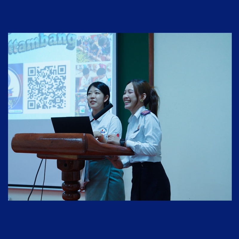 Two TukTuk staff in white uniform shirts present at a wooden lectern beside a screen showing a QR code