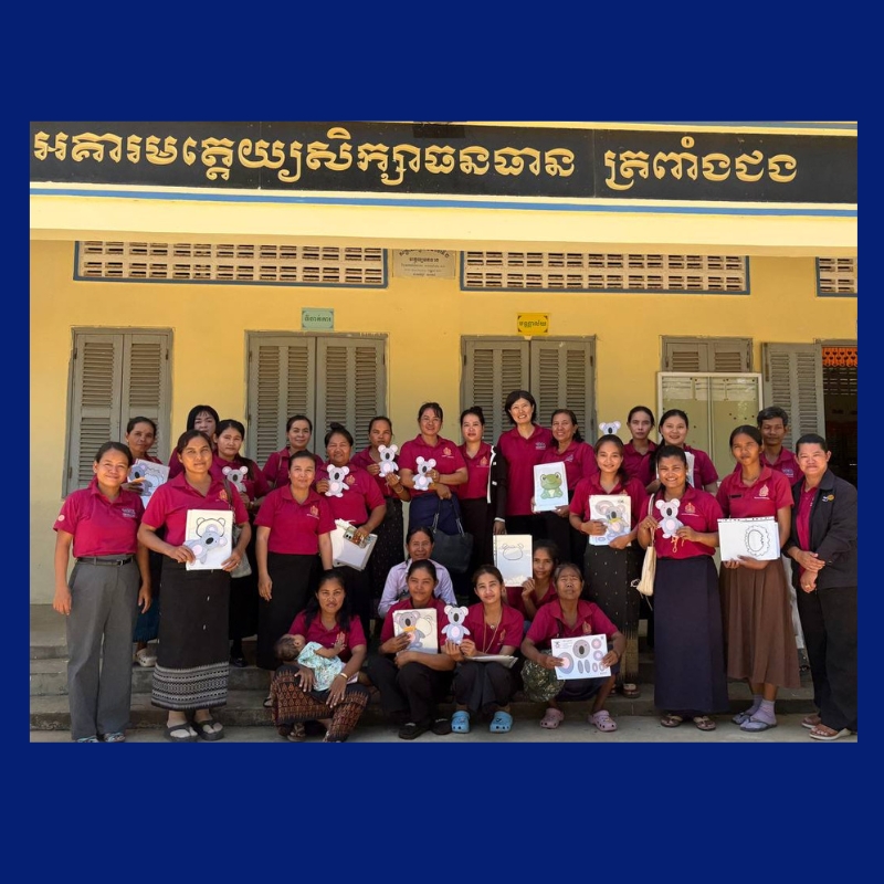 Teachers in maroon shirts and TukTuk staff pose outside a school holding paper animal crafts and worksheets