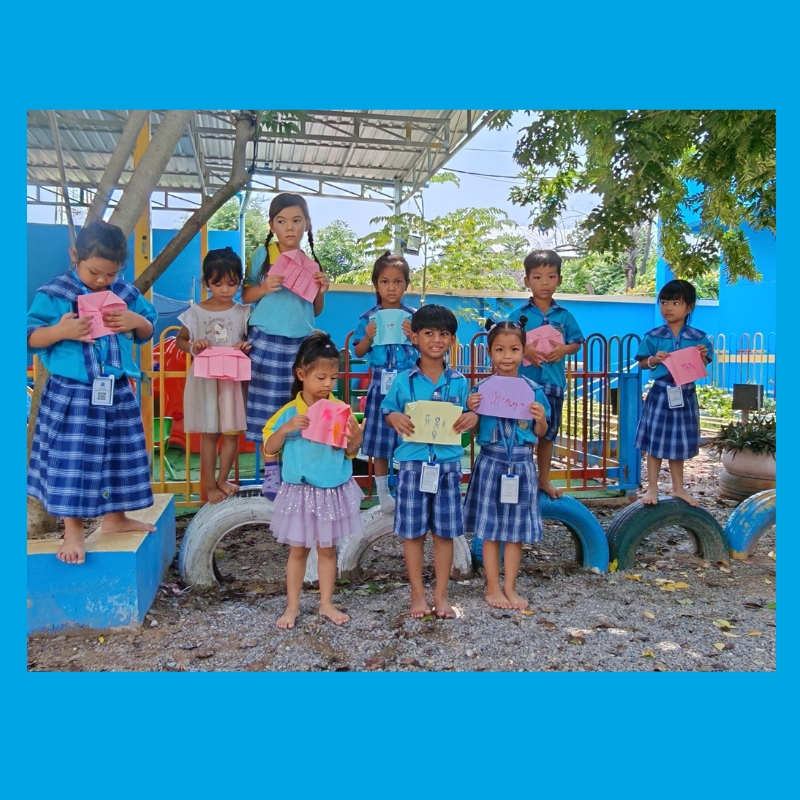 Preschool children in blue checked uniforms stand in a school playground holding folded pink and yellow paper crafts