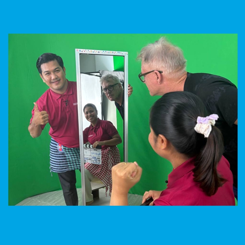 A TukTuk presenter gives a thumbs up beside a mirror on a green screen set while a filmmaker looks on