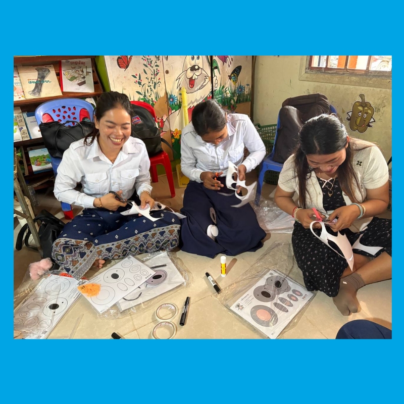 Three teachers sit on the floor cutting out paper masks and animal craft pieces during a workshop