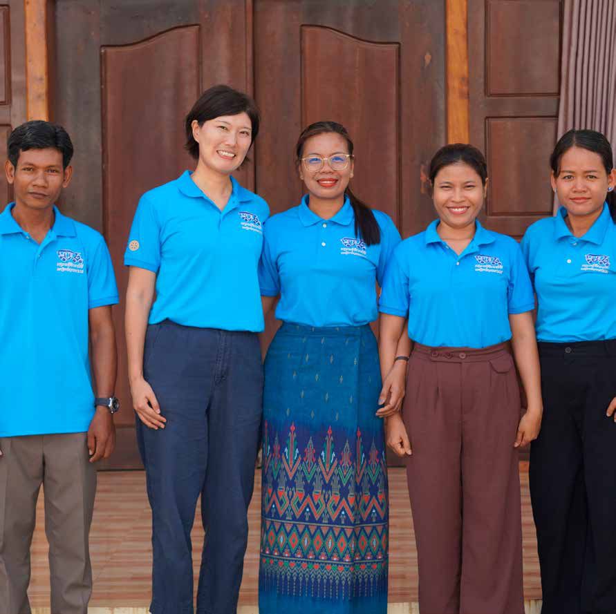 Five TukTuk staff and teachers in matching blue polo shirts smile together in front of a wooden door.