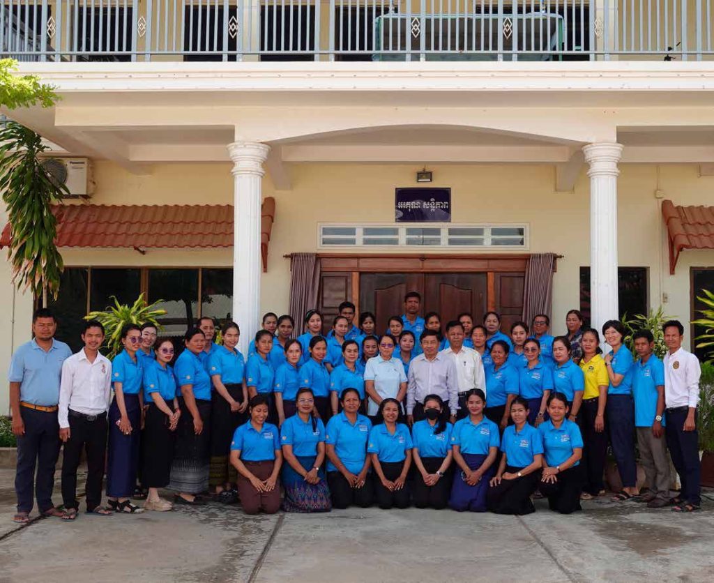 The TukTuk team and partner teachers in blue shirts pose together in front of a building in Cambodia.