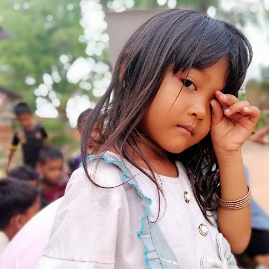 A young girl with long dark hair rubs her eye and looks at the camera outdoors in a village.